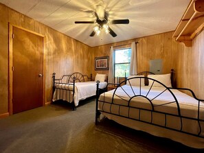 Ein Zimmer mit zwei Metallbetten, einem Deckenventilator, Holzwänden, einem Fenster und einer Tür.