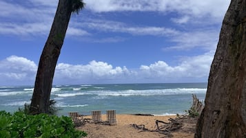 A beach with two wooden chairs and a sand area, surrounded by trees and ocean waves.