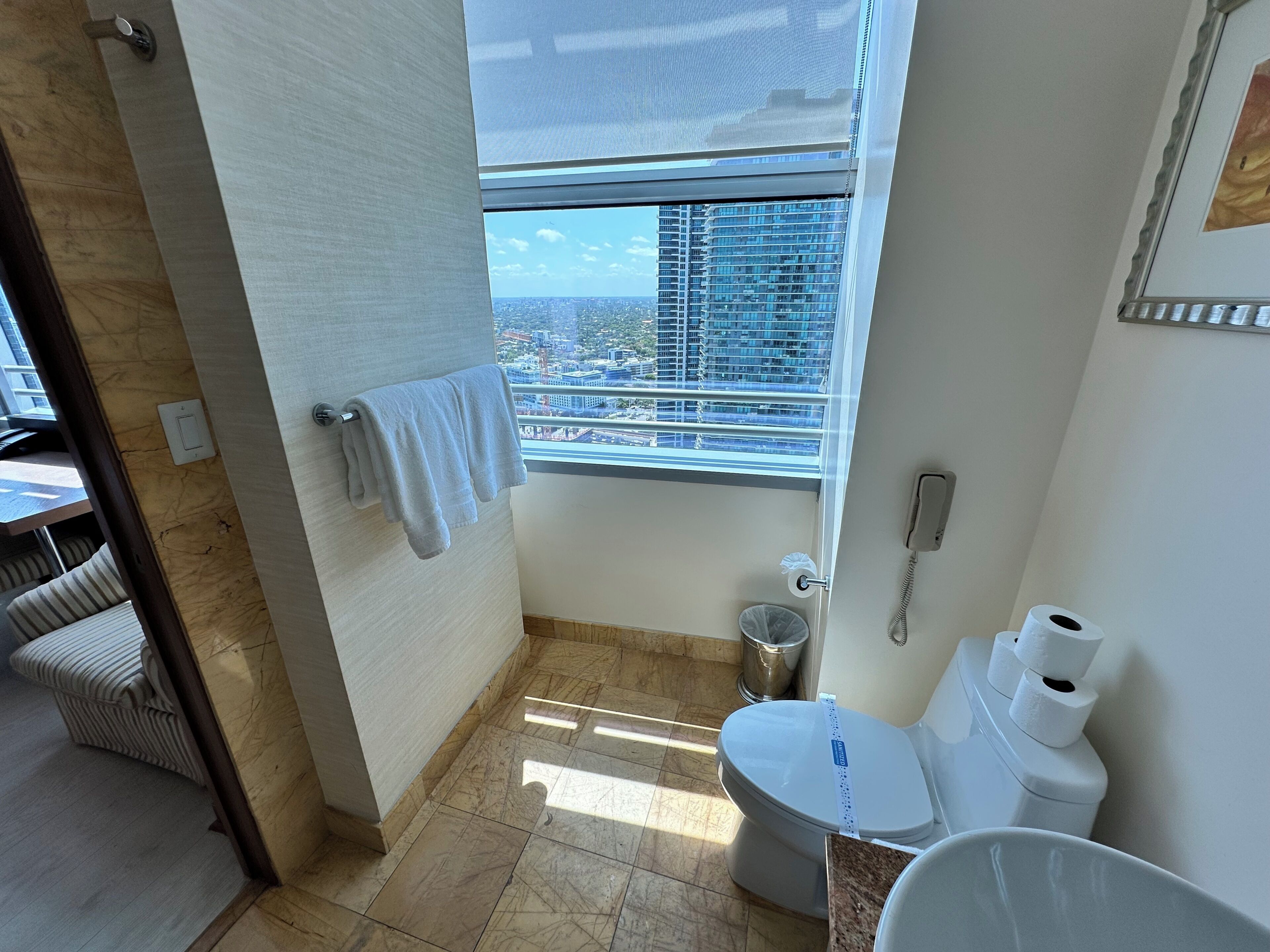 Une salle de bain équipée de toilettes, d’une poubelle, d’un porte-serviettes et d’une fenêtre donnant sur la ville.