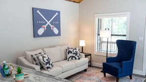 A living room with a sofa, a blue armchair, a coffee table, a lamp, and a window with blinds. - Picturesque Cabin Beautifully Designed in Livingston, Texas (Livingston)