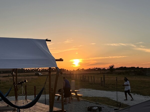 Une vue au coucher du soleil, avec une tente, un télescope et des personnes dans un champ d’herbe.