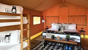 Habitación tipo tienda de campaña con literas, una cama central, un cartel de bienvenida, una lámpara y una pequeña mesa con libros y objetos decorativos.