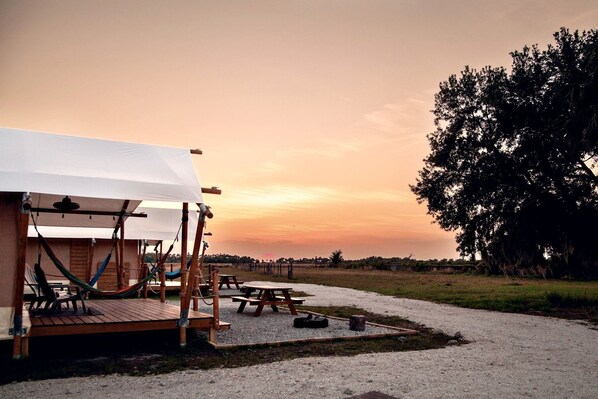 A rustic outdoor seating area with hammocks and a covered picnic table, set against a sunset backdrop.