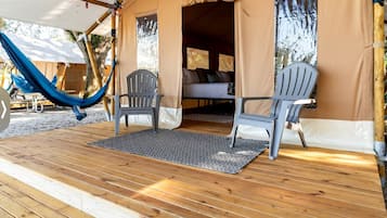 Een houten terras met twee stoelen en een hangmat, een overdekt buitengebied met een bed van binnenuit zichtbaar.