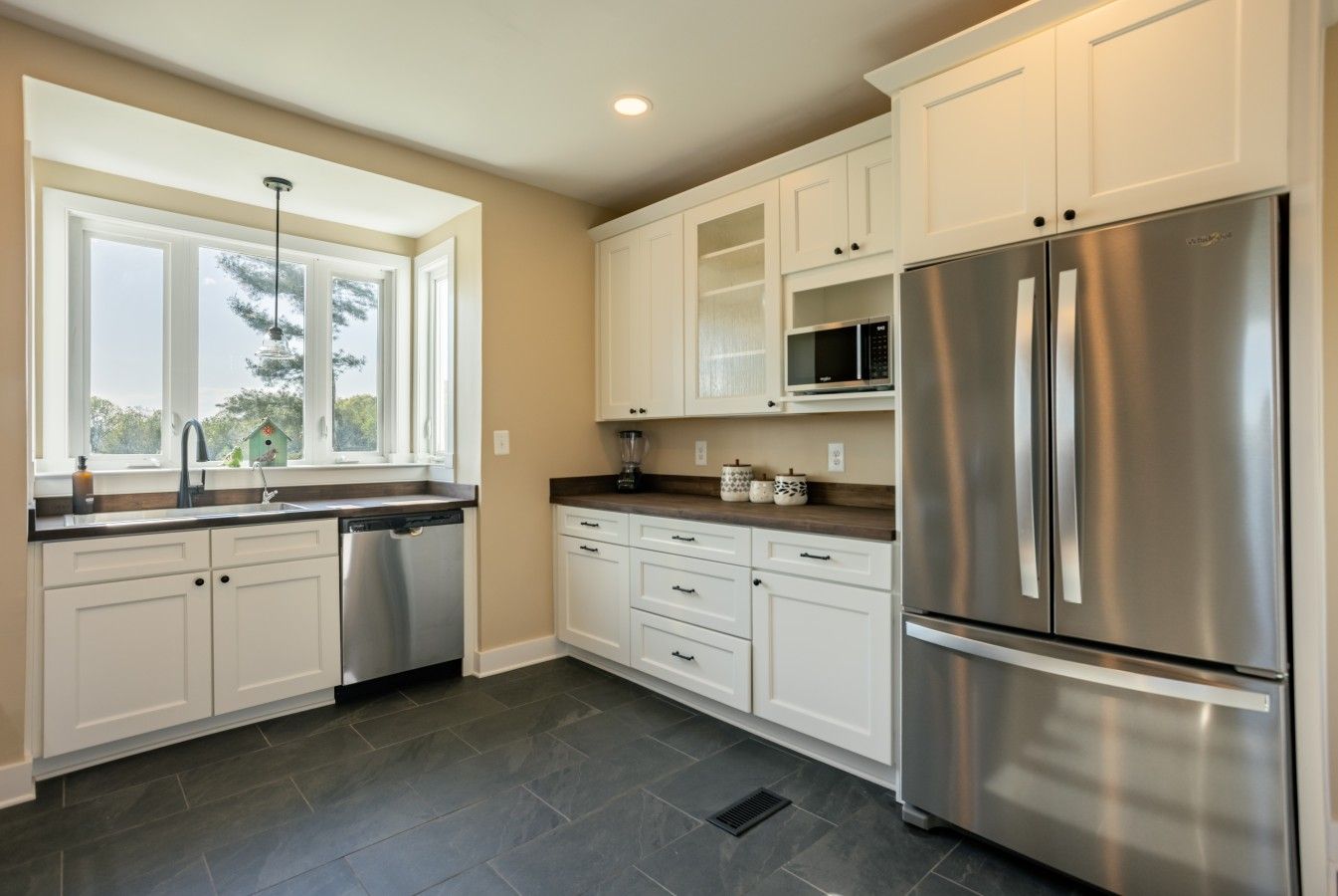 A kitchen with white cabinets, a stainless steel refrigerator, a dishwasher, and a window with a view of trees.