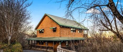 A log cabin with a green roof, a porch, and a stone wall in front.