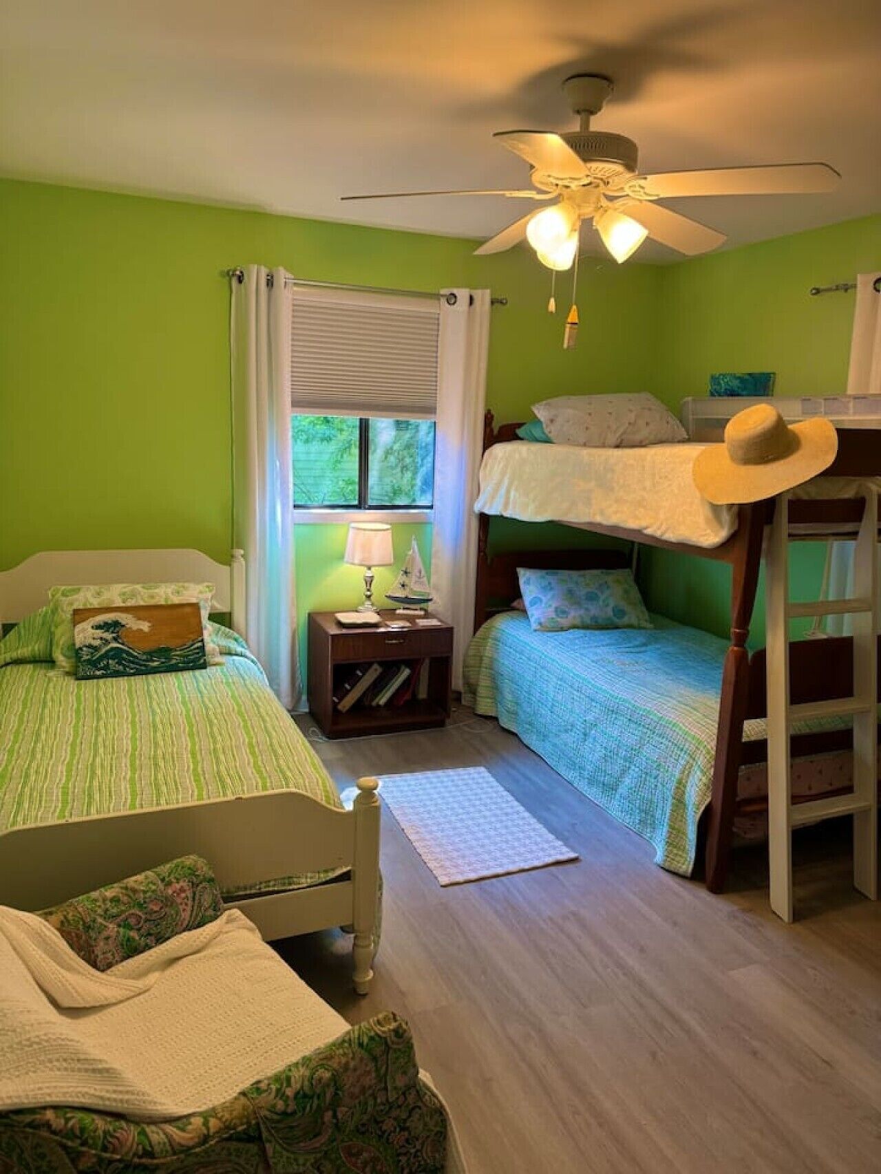 A room with two beds, a ceiling fan, a window with curtains, and a bedside table with a lamp.