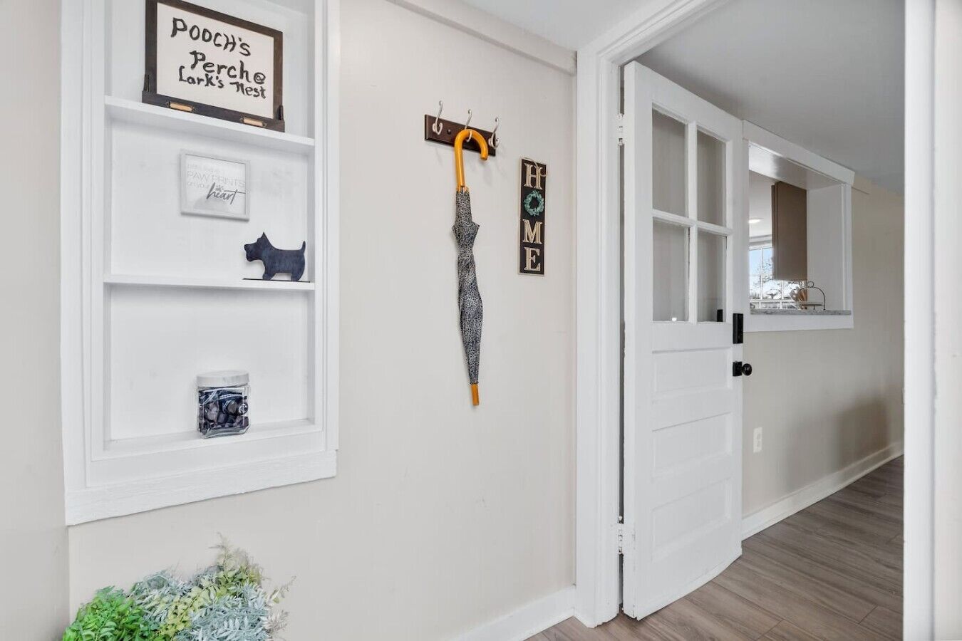 Un couloir avec une porte blanche, un porte-manteau avec un parapluie, une petite figurine de chien et une pancarte sur laquelle on peut lire « MAISON ».