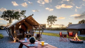 A rustic outdoor dining area with a wooden table set for two, a fire pit, and a tented structure with a dining table and chairs.