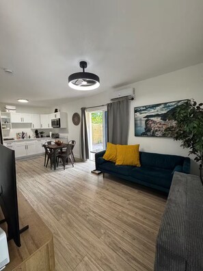 A modern living room with a dark blue sofa, a dining area with a wooden table, and a kitchen with white cabinets.