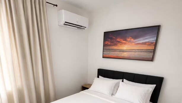 A neatly made bed with white linens and a folded towel, a black headboard, an air conditioning unit, and a sunset painting on the wall.