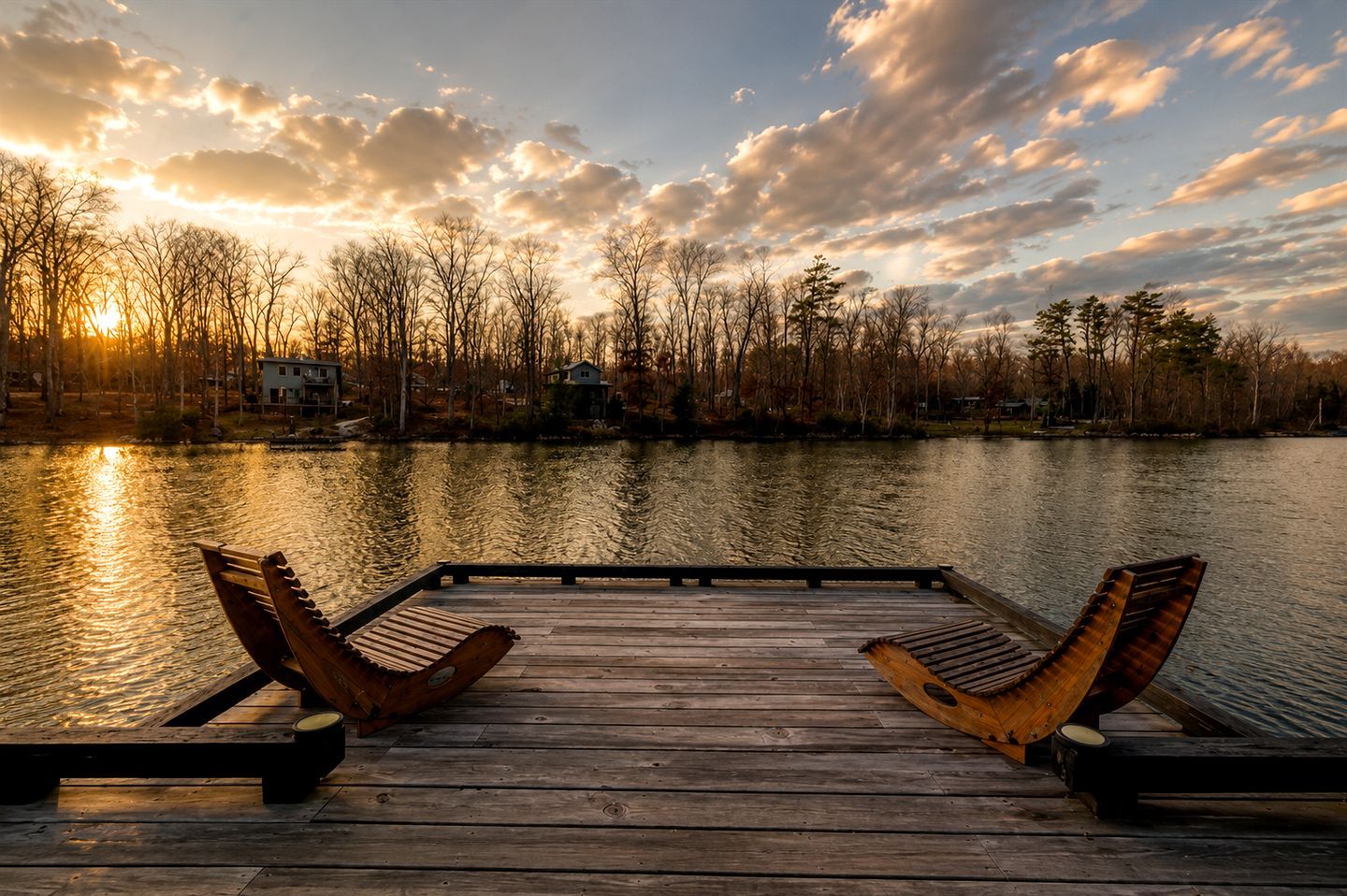 Un muelle de madera con dos sillas de madera mirando a un lago tranquilo, rodeado de árboles y casas al fondo.