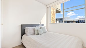 A bedroom with a bed, pillows, a window, and a view of buildings. - Tasman Towers - Unit 6 - Ocean Views (The Entrance)
