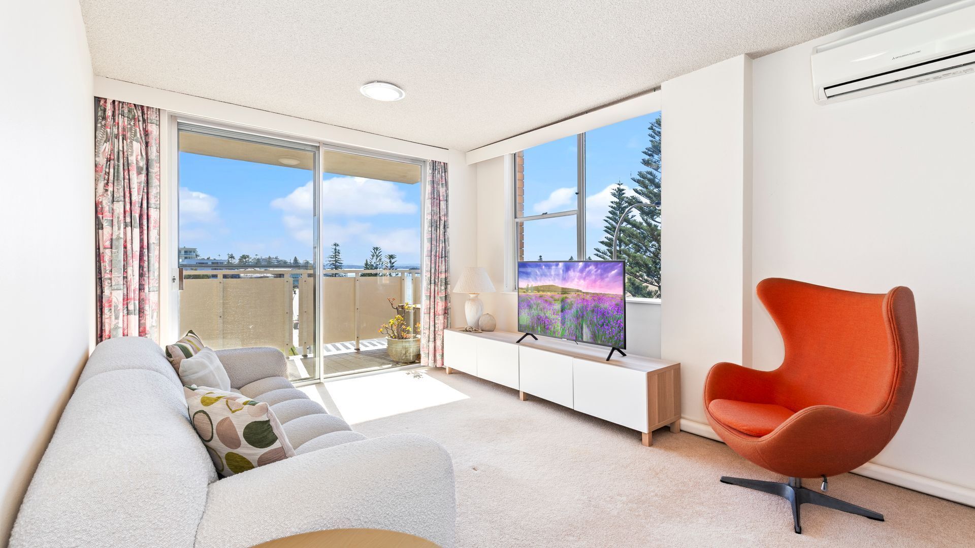 A modern living room with a sofa, a bright orange chair, a flat-screen TV, and a balcony with a view of the sky and trees.