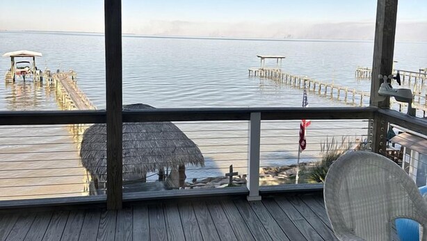 A wooden deck with a view of the sea, a thatched umbrella, and a wicker chair. - Luxury Bayfront Escape with Hot Tub & Fireplaces in Bacliff, Texas (Bacliff)