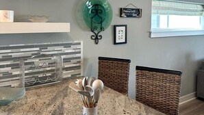 A kitchen with a granite countertop, hanging pendant lights, a green decorative plate, and wicker chairs. - Luxury Bayfront Escape with Hot Tub & Fireplaces in Bacliff, Texas (Bacliff)