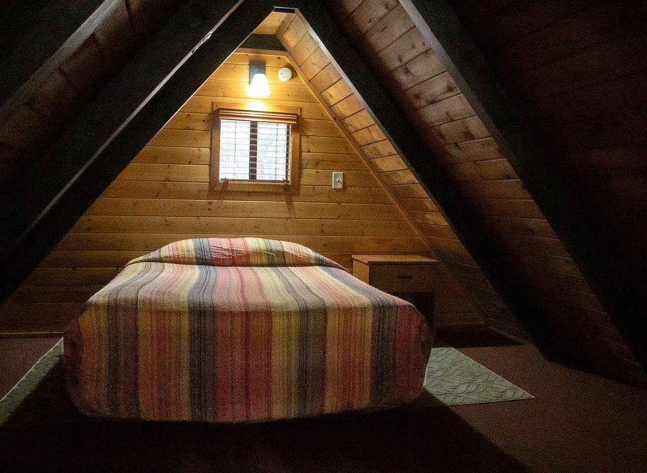Un dormitorio tipo loft con techo inclinado, una ventana con persianas, una cama con colcha a rayas y una mesita de noche pequeña.