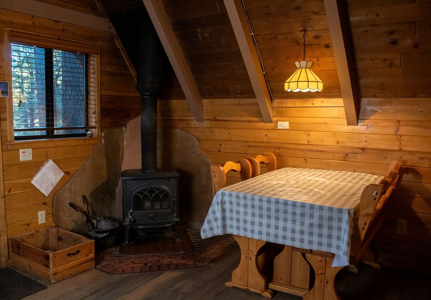 Interior acogedor de una cabaña con estufa de leña, una mesa con mantel a cuadros y una ventana con persianas.