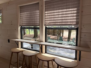 Ein Zimmer mit zwei Fenstern, jedes mit Blick auf Sitzgelegenheiten im Freien. Vor den Fenstern stehen drei hölzerne Hocker.