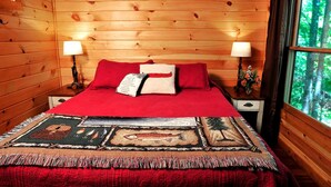 Une chambre dans une cabane en bois, avec un lit, deux lampes de chevet, une fenêtre donnant sur la forêt et un plaid à motifs.