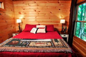 A wooden cabin bedroom with a bed, two bedside lamps, a window with a forest view, and a patterned throw blanket. - Tree House-Style Luxury Cabin in the Blue Ridge Mountains, North Carolina (Todd)