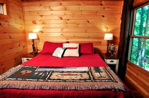 Ein Schlafzimmer in einer Holzhütte mit einem Bett, zwei Nachttischlampen, einem Fenster mit Blick auf den Wald und einer dekorativen Tagesdecke.