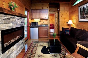 L’intérieur chaleureux d’un chalet avec une cheminée en pierre, des murs en bois, un coin cuisine et un salon comprenant un canapé et une lampe.