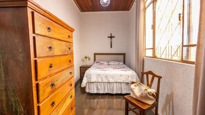 Ein Schlafzimmer mit einem hölzernen Schminktisch, einem Bett mit floralem Überwurf, einem Kruzifix an der Wand und einem Fenster mit Blick auf Gebäude.