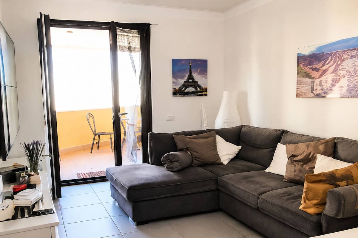 Una sala de estar con un sofá seccional gris, una mesita con lámpara y una puerta que da a un balcón.