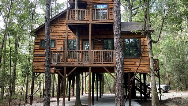 A wooden cabin with a balcony on the upper level, surrounded by trees. - Cozy Tree House with Beautiful Modern Interior in Livingston, Texas (Livingston)