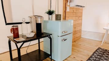 A compact kitchenette with a coffee maker, a small fridge, and a table with a mug and a bowl.