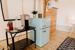A compact kitchenette with a coffee maker, a small fridge, and a table with a mug and a bowl.
