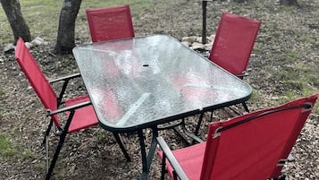 Una mesa cuadrada con cuatro sillas rojas en un entorno exterior, con árboles y un cobertizo al fondo.