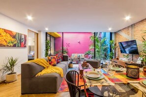 A modern living room with a sofa, dining table, and chairs. There is a TV mounted on the wall, and various plants are placed around the room.
