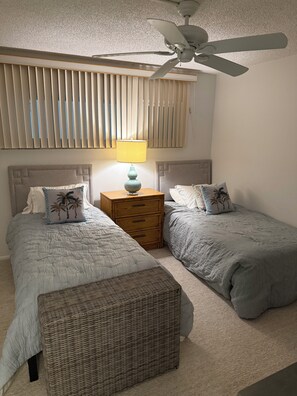 Een slaapkamer met twee bedden, een nachtkastje met lamp, een plafondventilator en een raam met zonwering.