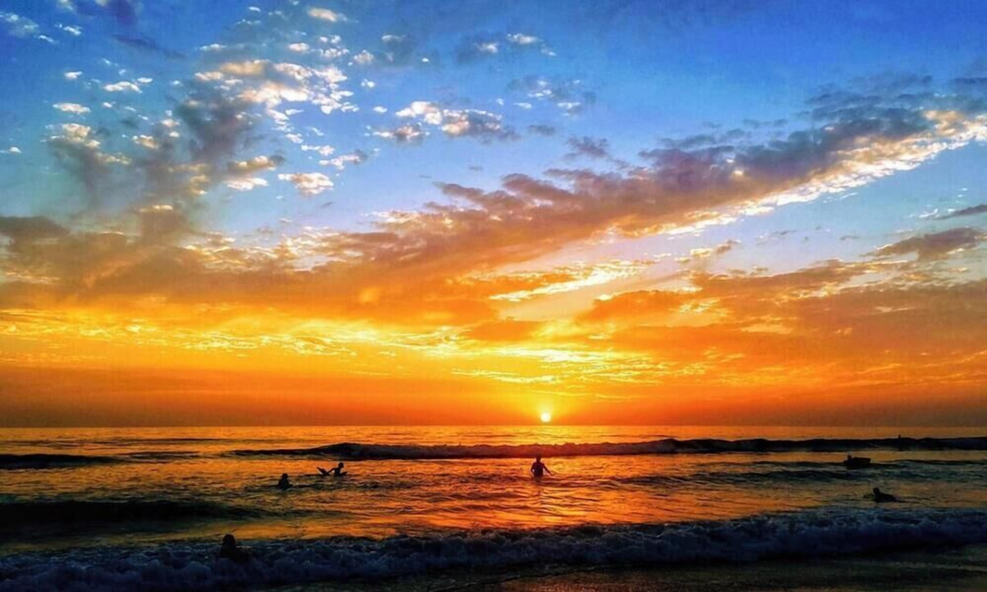 Un coucher de soleil sur l’océan, avec des vagues et les silhouettes de personnes.