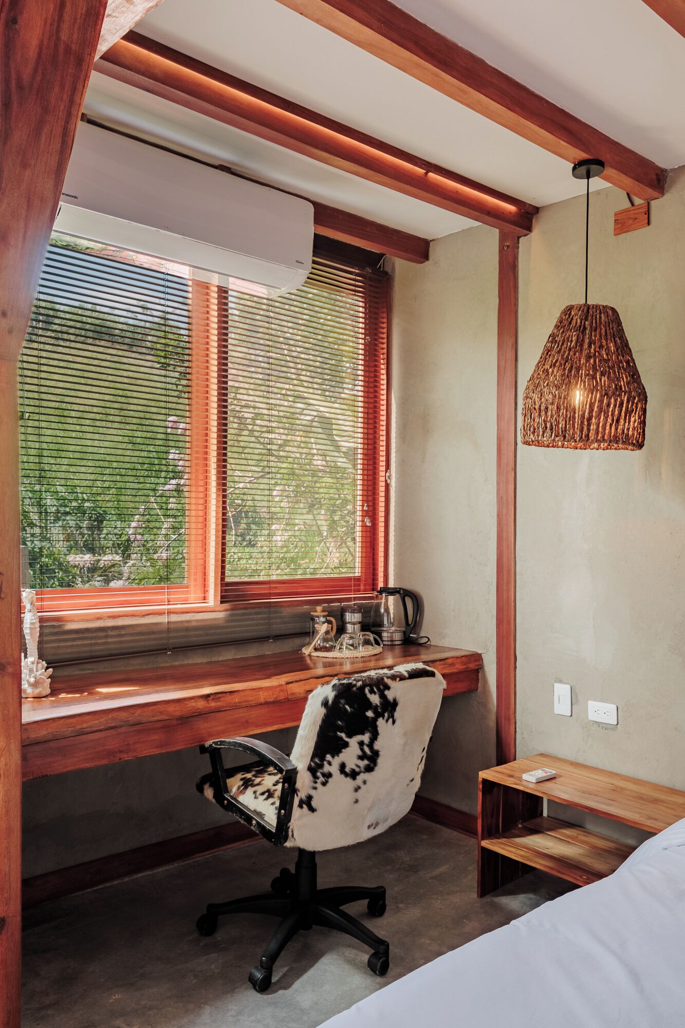 Un dormitorio con un escritorio de madera, una silla, una cama, una lámpara colgante y una ventana con persianas.