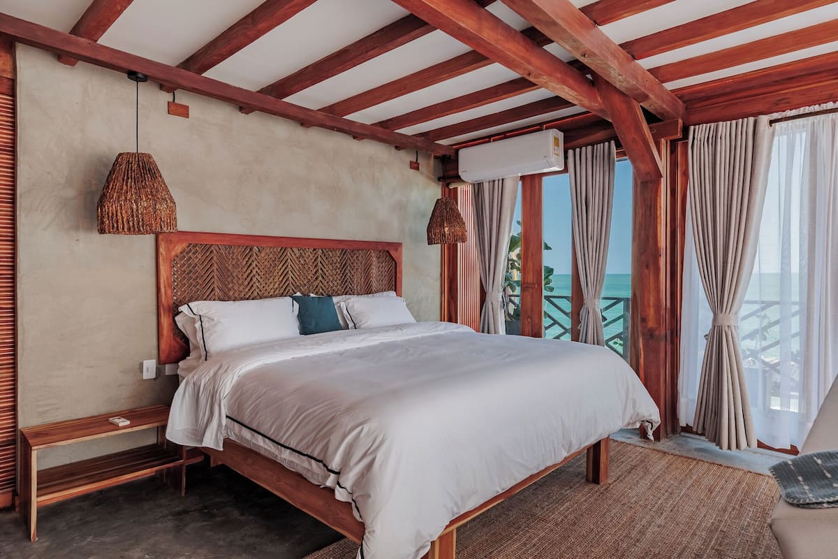 Un dormitorio con techo de madera, cama con ropa de cama blanca, lámpara tejida y vista al océano.