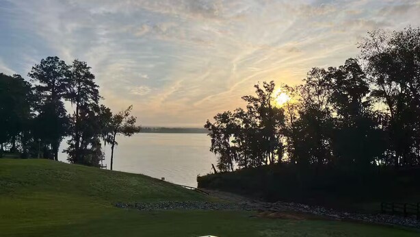 Näkymä järvelle auringonlaskun aikaan, taustalla puita ja ruohikkoista aluetta.