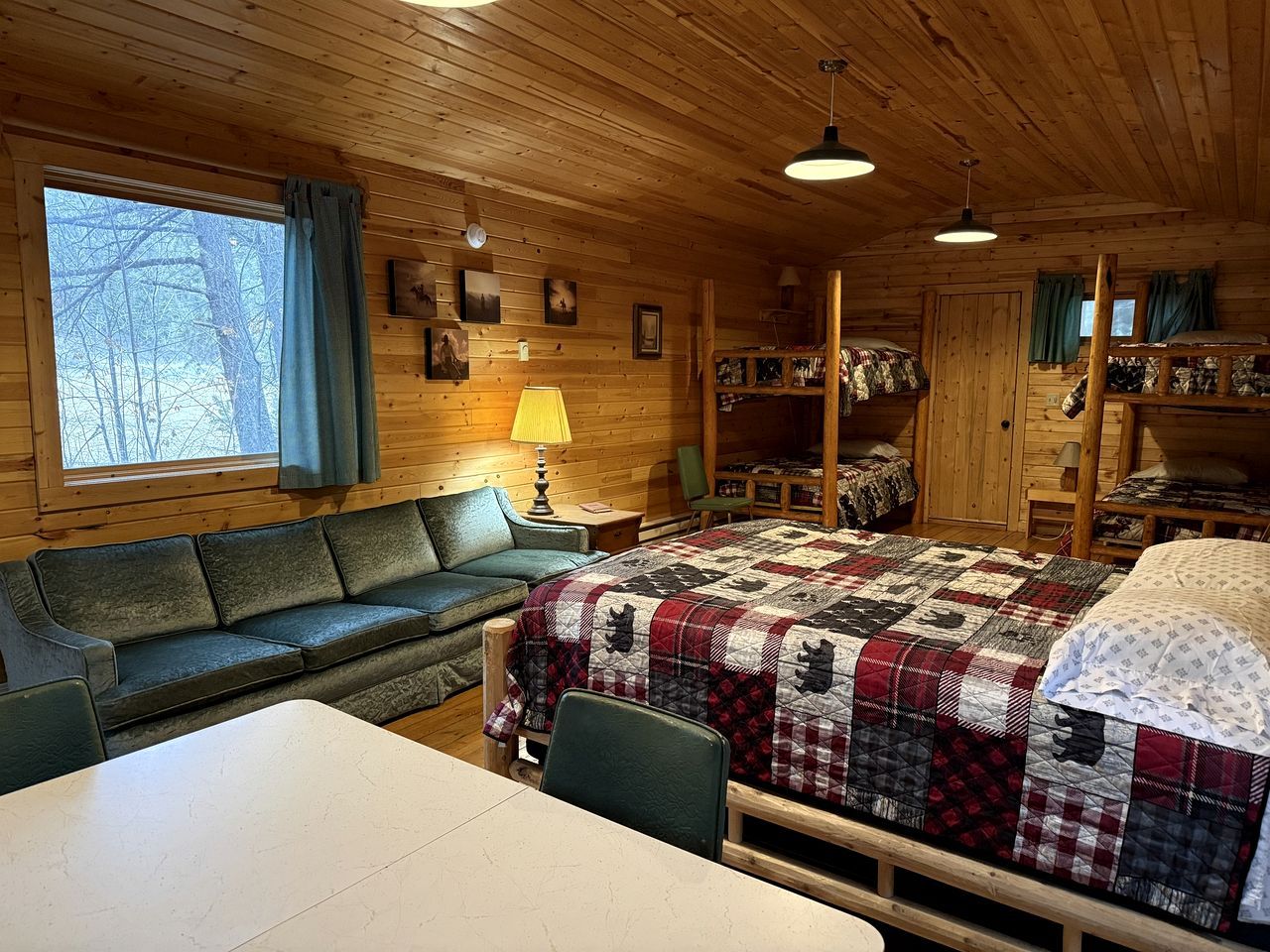 A cozy cabin interior with a large bed, a sofa, a dining table, and a window with curtains.