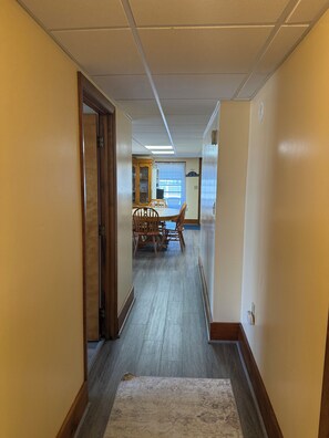 Um corredor com uma porta de madeira à esquerda, uma área de jantar com mesa e cadeiras no centro, e uma janela com persianas ao fundo.