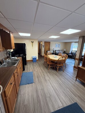 A kitchen with wooden cabinets, a black refrigerator, and a dining area with a table and chairs. - Beachfront 2-Bedroom Apartment on Wells Beach Sleeps 6 (Wells)