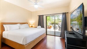 Quarto de hotel com cama, televisão, ventoinha no teto e porta de vidro deslizante para área externa com plantas.