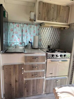 Une cuisine de taille réduite, avec des meubles en bois, une cuisinière à gaz, un évier et une fenêtre agrémentée de rideaux à motifs floraux.