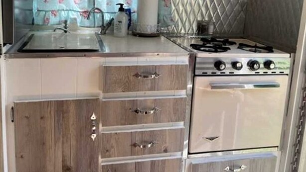 Une cuisine de taille réduite, avec des meubles en bois, une cuisinière à gaz, un évier et une fenêtre agrémentée de rideaux à motifs floraux.