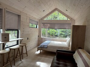 Interior de una cabaña con una cama, un escritorio con taburetes, una ventana con persianas y vista al bosque.