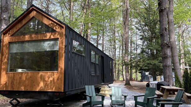 A small cabin with a wooden front and black siding, surrounded by trees. There are green Adirondack chairs and a picnic table outside.