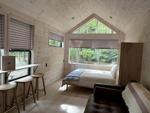 L’intérieur d’une cabane comprend un lit, un bureau avec des tabourets, une fenêtre avec des stores et une vue sur la forêt.