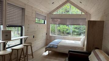 L’intérieur d’une cabane comprend un lit, un bureau avec des tabourets, une fenêtre avec des stores et une vue sur la forêt.