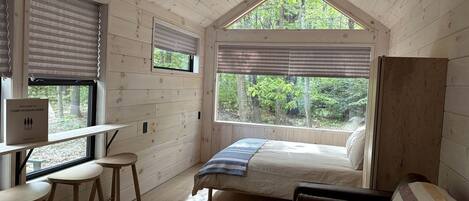 Ein Blockhaus-Interieur mit einem Bett, einem Schreibtisch mit Hockern, einem Fenster mit Jalousien und Blick auf den Wald.